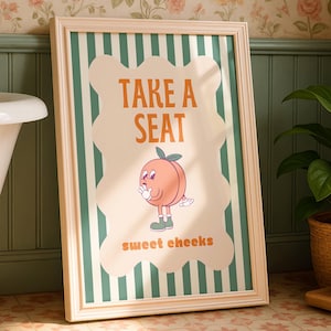 Puede incluir: Un cuadro enmarcado para el baño con el texto "TAKE A SEAT" y "sweet cheeks". La obra presenta un personaje de melocotón de dibujos animados. El fondo tiene rayas verticales verdes y blancas.