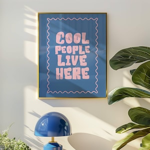 May include: A framed art print with the text "COOL PEOPLE LIVE HERE" in pink letters on a blue background. A blue table lamp with a gold base sits on a white surface. A potted plant is in the foreground.