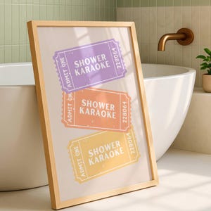 May include: A framed print featuring three colorful tickets with the text "Shower Karaoke." The tickets are in purple, orange, and yellow, with the words "Admit One" and a unique number. The frame is made of light wood, and the print is displayed in a bathroom setting.