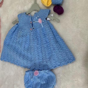 May include: A light blue crochet baby dress and matching diaper cover set. The dress has a scalloped hem and is adorned with pink flower accents. Three balls of yarn in yellow, blue, and purple are visible.