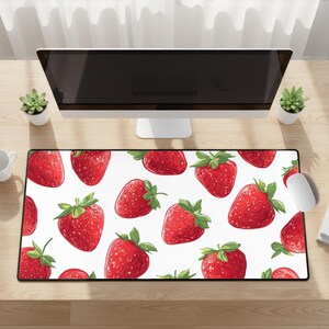 Può includere: Tappetino per mouse da scrivania rettangolare con un motivo di fragole rosso vivo con foglie verdi su sfondo bianco. Il tappetino è su una scrivania in legno chiaro, con un monitor del computer, un mouse e piccole piante visibili.