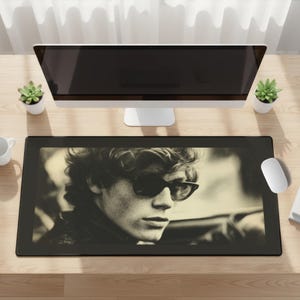 May include: Black rectangular desk mat featuring a sepia-toned image of a person wearing sunglasses. The desk mat is placed on a light-colored wooden desk, with a computer monitor, succulents, and a mouse visible.