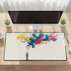 May include: A rectangular desk mat with a black border and a cream-colored surface. The mat features a whimsical illustration of a person with a thought bubble of colorful paint splatters in yellow, blue, and pink. A computer monitor, mouse, and plants are on the desk.