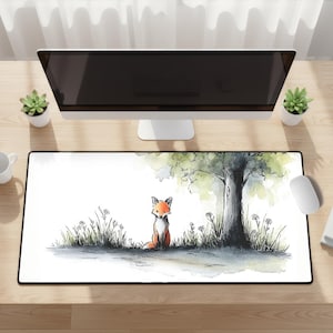 May include: A rectangular desk mat featuring a watercolor illustration of a small orange and white fox sitting in grass near a tree. The artwork uses black ink outlines and soft green and yellow washes on a white background, creating a nature scene.