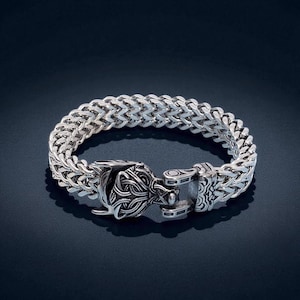 May include: A silver-colored bracelet with a woven chain design. The bracelet features a detailed clasp with an ornate, stylized animal head. The metal bracelet is displayed on a dark blue background.