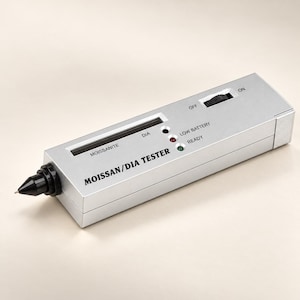 May include: A silver-colored handheld Moissan/Dia tester device with a black probe tip. The unit features an on/off switch, indicator lights for Moissanite, Dia, low battery, and ready status, with the text MOISSAN/DIA TESTER printed on the front.