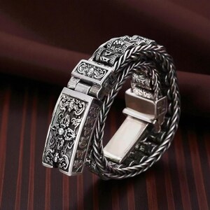 May include: A silver-colored bracelet with an intricate design. The bracelet features a braided band and rectangular clasps with detailed carvings. The clasps have a decorative pattern and engraved text. The bracelet is displayed on a dark brown surface.