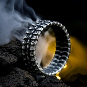 Peut inclure: Une bague argentée avec un motif texturé en forme d'écailles. L'anneau est présenté sur un fond sombre avec des volutes de fumée, mettant en valeur ses détails complexes. Le design de la bague rappelle les écailles de dragon, un bijou unique.
