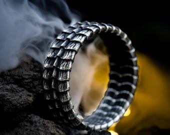May include: A silver-colored ring with a textured, scale-like design. The ring is set against a dark background with a hint of smoke, highlighting its intricate details. The ring's design resembles dragon scales, making it a unique piece of jewelry.