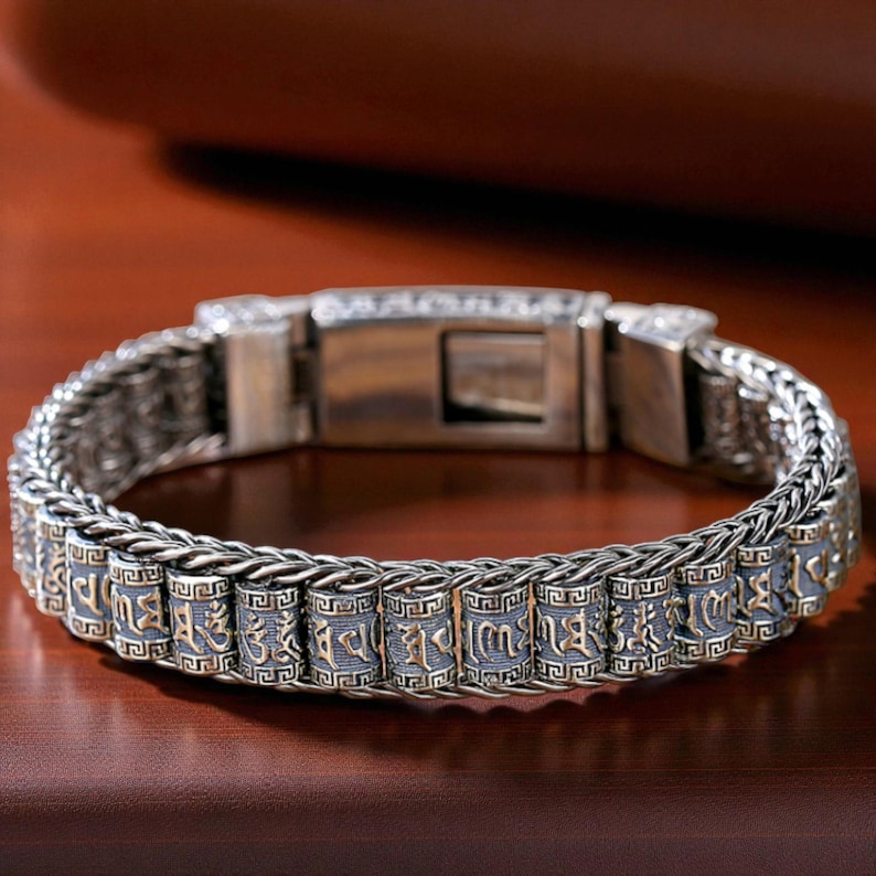 May include: Silver-toned bracelet with a series of cylindrical beads inscribed with gold-colored symbols. The bracelet features a woven chain design and a secure clasp. The beads are accented with a geometric pattern.