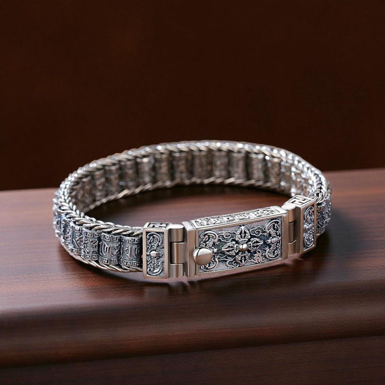 May include: A silver bracelet with an intricate design. The bracelet features a woven chain and a rectangular clasp with detailed patterns. The clasp has a central button and is flanked by decorative elements. The bracelet is displayed on a wooden surface.