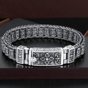 May include: A silver-colored bracelet with intricate engraved patterns. The bracelet features a central rectangular clasp with detailed designs and smaller, cylindrical links with script. The design is ornate and detailed.