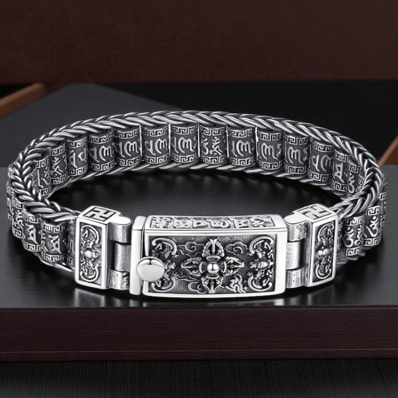 May include: A silver-colored bracelet with intricate engraved patterns. The bracelet features a central rectangular clasp with detailed designs and smaller, cylindrical links with script. The design is ornate and detailed.
