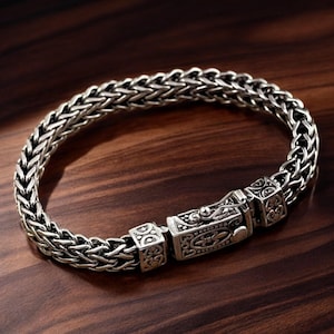 Peut inclure: Bracelet argenté avec un motif de chaîne tressée. Le bracelet est doté d'un fermoir orné de détails gravés. Le bijou est présenté sur une surface en bois sombre, soulignant l'éclat métallique du bracelet.