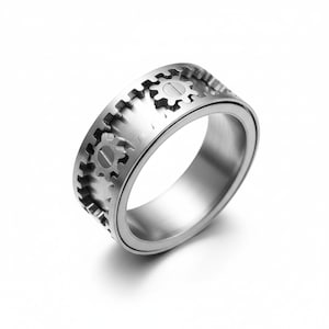 May include: A silver-colored ring with a gear design. The ring features a series of small gear shapes around the band. The gears are etched into the metal, creating a detailed and mechanical aesthetic. The ring has a smooth, polished finish.