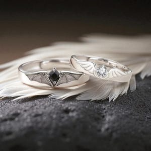 Puede incluir: Dos anillos de plata, uno con una gema negra cuadrada y diseño de alas de murciélago, el otro con una gema redonda transparente y diseño de alas de ángel. Los anillos se exhiben sobre una pluma blanca.