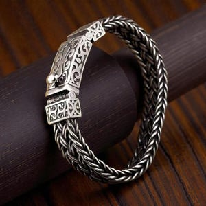 Pode incluir: Uma pulseira de prata com um design de corrente trançada. O fecho apresenta intrincados arabescos e um fecho seguro. A pulseira é feita de um material metálico brilhante e é exibida em uma superfície de madeira escura.