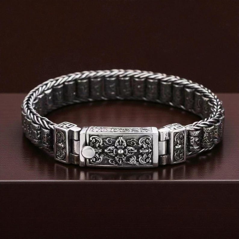 May include: A silver-colored bracelet with an intricate design. The bracelet features a woven chain and a rectangular clasp with detailed carvings. The clasp has a small, round button. The bracelet is displayed on a dark brown surface.