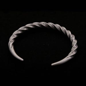 May include: A silver-colored cuff bracelet with a twisted rope design. The open-ended bracelet has a textured surface and is set against a black background. The bracelet is a piece of jewelry.