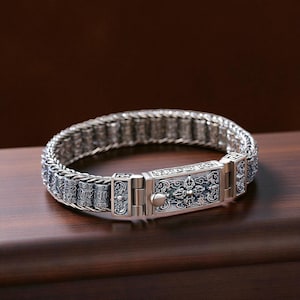 May include: A silver bracelet with intricate detailing. The bracelet features a woven chain design and a rectangular clasp with ornate patterns. The clasp has a central button and is flanked by decorative elements. The bracelet is displayed on a wooden surface.