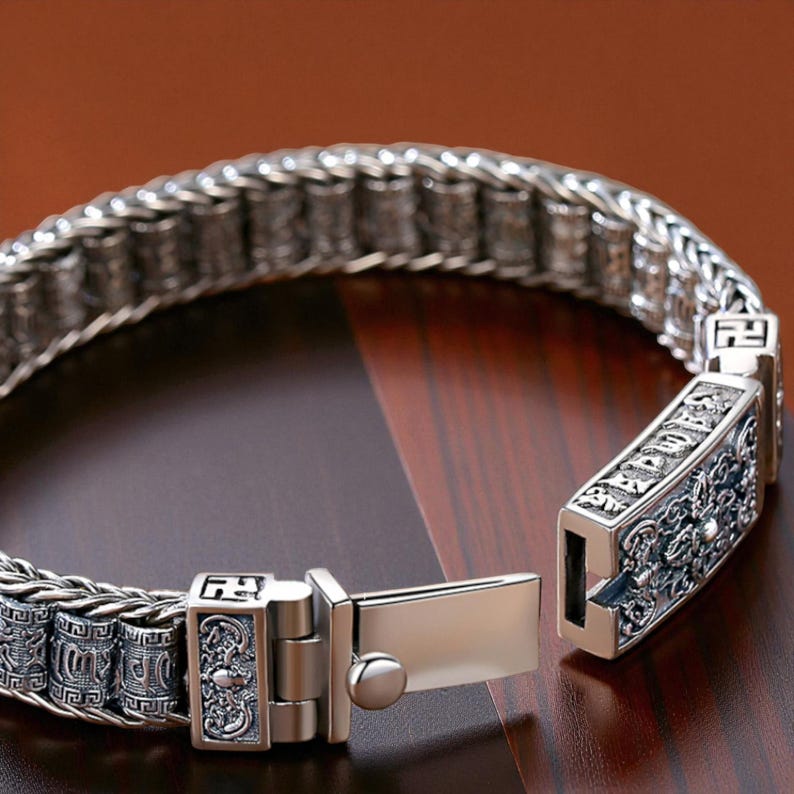May include: Close-up of a silver bracelet with an intricate chain design. The bracelet features detailed decorative clasps with engraved patterns. The clasp is open, revealing the inner workings. The bracelet is displayed on a dark wooden surface.
