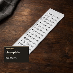 May include: A rectangular tungsten carbide drawplate for round wire, featuring multiple precision-drilled holes with sizes ranging from 0.26 mm to 4.10 mm clearly engraved next to each opening. The metal tool has a brushed finish and text.