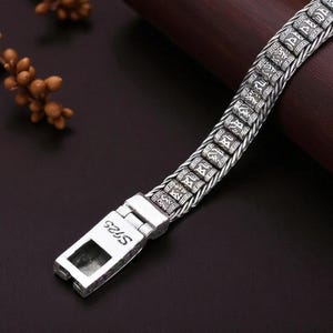 May include: Close-up of a silver bracelet with an intricate design. The bracelet features rectangular links with engraved patterns and text, connected by a braided chain. The clasp is marked with "S925".