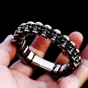 May include: A silver-colored bracelet featuring a series of skull-shaped links. The skulls are detailed with eye sockets, noses, and teeth. The bracelet is held in a hand against a dark background, showcasing its metallic sheen.