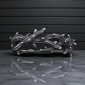 May include: A silver-toned metal bracelet with a thorny branch design. The bracelet has a textured surface and is reflected in the surface below. The bracelet is a unique piece of jewelry.