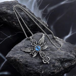 May include: Silver-toned pendant necklace featuring a cross design with ornate detailing and small wings. The center of the cross has a blue eye-shaped stone. The necklace is displayed on a dark, textured rock.