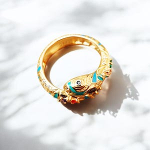 Può includere: Un anello dorato con un design a forma di pesce. L'anello presenta dettagli in smalto turchese, rosso, giallo e viola. Il pesce ha un occhio nero ed è circondato da piccole pietre preziose trasparenti. L'anello è su uno sfondo di marmo bianco.