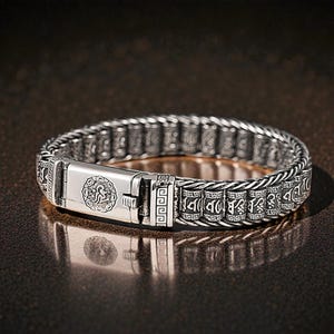 Puede incluir: Pulsera plateada con un diseño intrincado. La pulsera presenta una serie de eslabones rectangulares con patrones grabados y un cierre central con un emblema circular. El metal tiene una apariencia ligeramente envejecida, realzando los detalles.