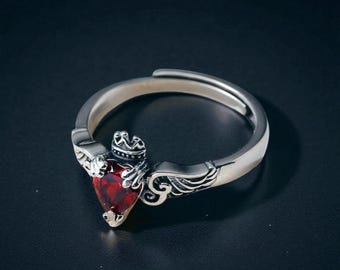 Peut inclure: Bague en argent ornée d'une pierre rouge foncé en forme de cœur. La bague présente une petite couronne au-dessus du cœur et des détails décoratifs d'ailes et de volutes sur les côtés. La bague est posée sur un fond sombre.