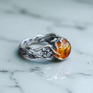 Puede incluir: Un anillo de plata con una piedra grande y redonda de color ámbar. La banda del anillo tiene un diseño texturizado en forma de rama con un pequeño detalle floral. La piedra de ámbar es el punto focal, engastada en un engaste detallado inspirado en la naturaleza.