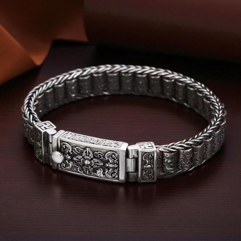 May include: A silver-colored bracelet with an intricate design. The bracelet features a woven chain design with detailed rectangular clasps and decorative elements. The central clasp has an ornate pattern, and the bracelet is displayed on a dark surface.
