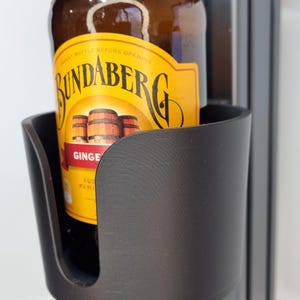 May include: A black 3D-printed cup holder containing a brown glass bottle of Bundaberg Ginger Beer. The bottle has a yellow label with red text and a logo of three barrels. The cup holder is mounted on a black metal frame.