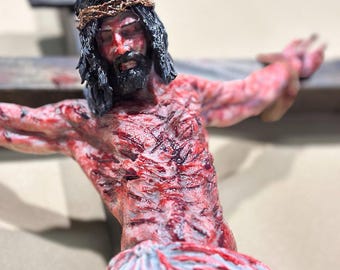 May include: A detailed sculpture of Jesus Christ on a wooden cross. The figure has dark hair, a beard, and a crown of thorns. The body is painted with red and white hues, depicting wounds. The cross is weathered and gray.
