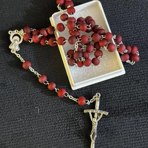 Puede incluir: Un rosario con cuentas de color rojo intenso y una cadena y crucifijo de metal plateado. Las cuentas tienen un acabado mate, y el crucifijo presenta una detallada representación de Jesús. El rosario está parcialmente dentro de una caja blanca.