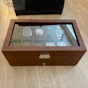 May include: A rectangular brown watch storage box with a glass lid and a silver metal latch. The interior features twelve individual compartments, with six watches currently displayed inside. A small drawer is located at the bottom with a silver knob.