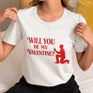 以下が含まれることがあります： 赤い文字で「WILL YOU BE MY VALENTINE？」と書かれた白いTシャツ。その下に、ひざまずいて指輪を持っている人の赤いシルエットがプリントされています。カジュアルなスタイルのTシャツです。