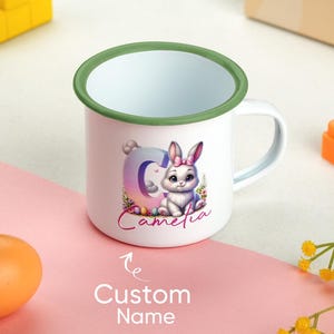 Könnte beinhalten: Weißer Emaille-Becher mit grünem Rand und Henkel. Der Becher zeigt ein Cartoon-Kaninchen mit rosa Schleife, den Buchstaben "C" und den Namen "Camelia". Darunter steht "Custom Name".