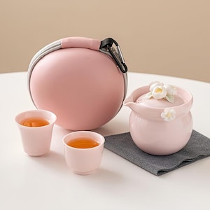 Puede incluir: Un juego de té rosa con una tetera, dos tazas y un estuche. La tetera tiene un diseño floral en la tapa. Las tazas y la tetera están llenas de té. El estuche es redondo y tiene un asa.