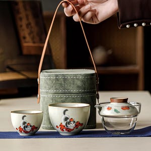 May include: A tea set featuring two ceramic cups and a glass teapot with a lid, all decorated with illustrations of a cat and orange fruits. The set includes a cylindrical green carrying case with a tan leather handle for transport.