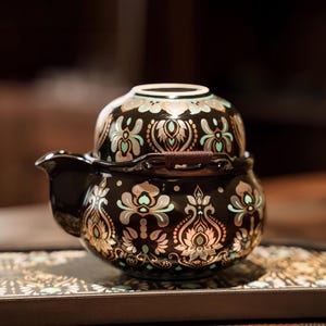 May include: A black ceramic teapot with a matching cup stacked on top, featuring an intricate repeating pattern in metallic gold and soft mint green. The teapot has a curved spout and a small brown woven handle detail on the lid rim.