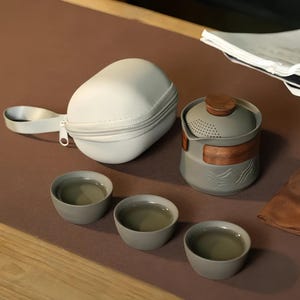 Könnte beinhalten: Ein graues Teeservice mit Holzdeckel und Akzenten, einschließlich einer Teekanne, drei kleinen Tassen und einer Tragetasche. Die Teekanne hat ein Bergdesign. Die Tassen und die Teekanne sind mit Tee gefüllt.