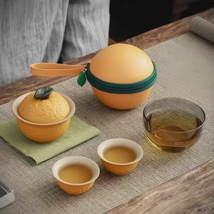 Puede incluir: Un juego de té con temática de cítricos. El juego incluye un recipiente redondo naranja con cremallera verde, un cuenco pequeño con tapa, dos tazas de té pequeñas y un vaso transparente. Las tazas y el cuenco están llenos de té.