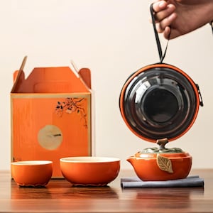 Puede incluir: Un juego de té de cerámica naranja con una tetera, dos tazas y un estuche. La tetera tiene un asa en forma de hoja y una hoja decorativa. El juego se exhibe con una caja de regalo naranja.