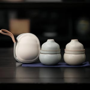 Może przedstawiać: Ceramiczny zestaw do herbaty z dwoma filiżankami z pokrywkami i etui. Filiżanki są jasnoniebieskie i białe, z brązową obwódką. Etui jest białe z szarą lamówką i beżowym uchwytem.