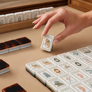 Puede incluir: Un juego de fichas de mahjong rectangulares con caras blancas que presentan diversas ilustraciones, incluyendo un leopardo, pagodas y diseños florales, cada una etiquetada con la palabra JOKER. Las fichas tienen bases en capas de color marrón oscuro, azul claro y blanco.