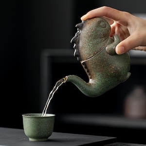 Puede incluir: Una tetera de cerámica texturizada en verde y marrón con forma de criatura, con un hocico y una cresta dorsal prominentes, vertiendo líquido claro en una pequeña taza de cerámica verde a juego. La tetera tiene un acabado moteado.